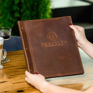 Może przedstawiać: Brązowe skórzane menu z wytłoczonym napisem "TANTALUM" na okładce. Menu jest otwarte, ukazując wewnętrzne strony. Na drewnianym stole stoi latte i szklanka ciemnego płynu.