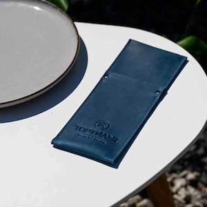 Puede incluir: Un portamenús de cuero azul oscuro con el texto en relieve "TOPИAЛЬ" y un logotipo, apoyado sobre una mesa blanca junto a un plato gris con borde marrón. La escena está iluminada por luz natural, con vegetación en el fondo.