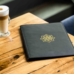 Puede incluir: Un café con leche en un vaso transparente con asa está sobre una mesa de madera junto a un menú de cuero negro con un diseño floral dorado. La mesa tiene vetas de madera natural.