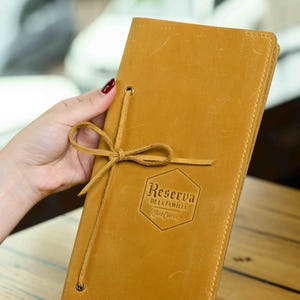 Peut inclure: Une couverture de menu en cuir de couleur moutarde avec une fermeture à nœud. La couverture porte le texte en relief "Reserva de la Familia" dans un cadre hexagonal. Le menu est fermé et tenu par une main.