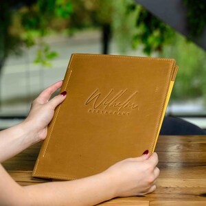 May include: A tan leather menu with the embossed text "Wilhelm Restaurant" is held open. The menu has a stitched border and a yellow interior. The background is blurred, with a wooden table and greenery visible.