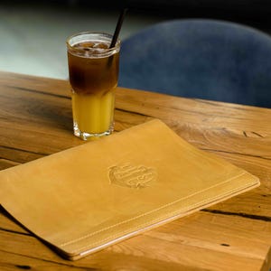 May include: A layered drink in a tall glass with a straw sits on a wooden table next to a tan leather menu. The drink has a yellow base and a dark brown top. The menu has an embossed design.