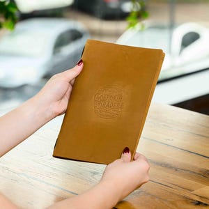 Op de afbeelding: Een bruin leren menu met de tekst "Goldforth Traders" in reliëf wordt vastgehouden. Het menu is rechthoekig en gesloten. Een glas latte staat op een houten tafel.