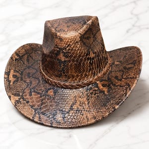 May include: A brown and black cowboy hat featuring a detailed snakeskin pattern across the entire surface. The hat includes a pinched crown and a wide, curved brim, accented by a braided band that encircles the base of the crown.