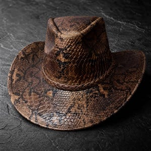 May include: A brown and black cowboy hat featuring a detailed snakeskin pattern across the crown and brim. The hat includes a braided band encircling the base of the crown, with a pinched top design and a wide, slightly curved brim.