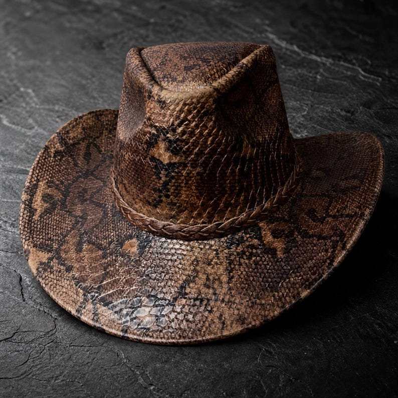 May include: A brown and black cowboy hat featuring a detailed snakeskin pattern across the crown and brim. The hat includes a braided band encircling the base of the crown, with a pinched top design and a wide, slightly curved brim.