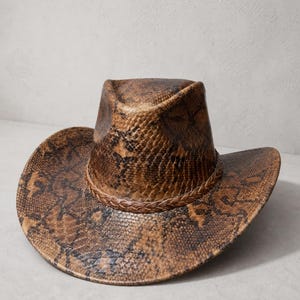 May include: A cowboy hat featuring a brown and black snakeskin pattern throughout the entire surface. The hat has a pinched crown and a wide, curved brim, accented by a braided brown band encircling the base of the crown.