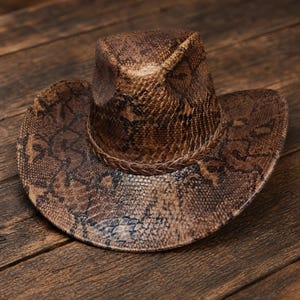 May include: A cowboy hat featuring a brown and black snakeskin pattern throughout the entire surface. The hat has a pinched crown and a wide, curved brim, accented by a braided band that encircles the base of the crown for a textured finish.