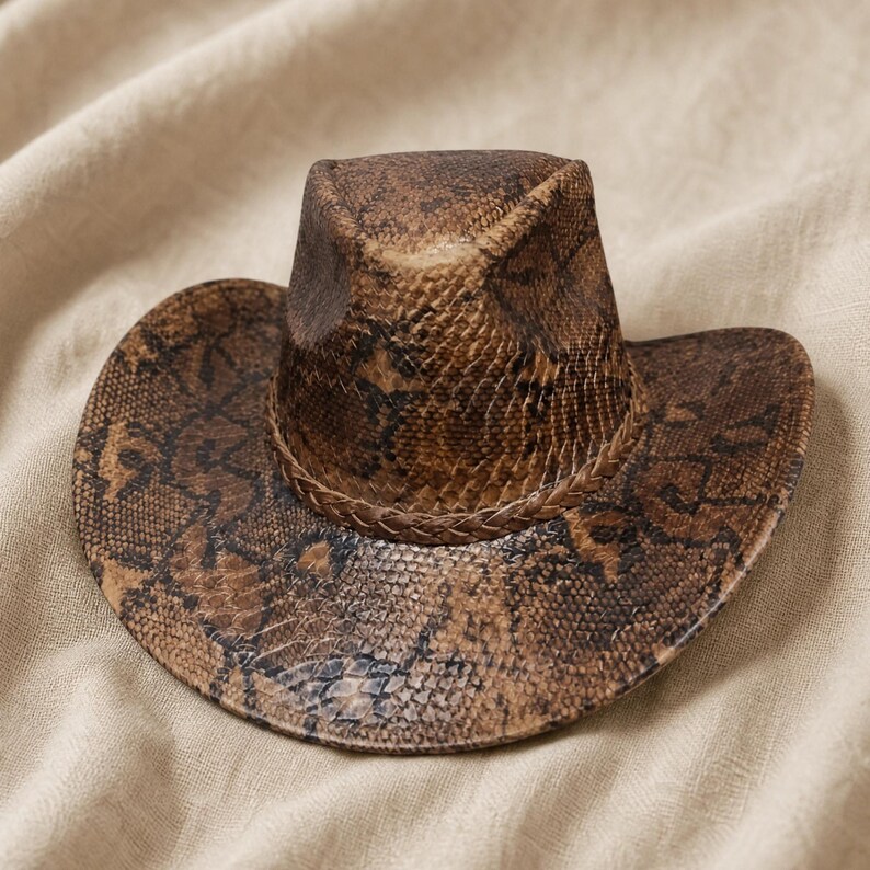 May include: A brown and black snakeskin patterned cowboy hat featuring a pinched crown and a wide, curved brim. The hat is accented with a braided band that encircles the base of the crown, matching the overall textured reptilian print design.