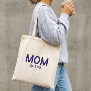 Puede incluir: Un bolso de tela color crema con el texto azul oscuro "MOM OF TWO" impreso en la parte delantera. El bolso tiene dos correas largas para el hombro y una forma rectangular, confeccionado con un material de tela tejida con costuras visibles.