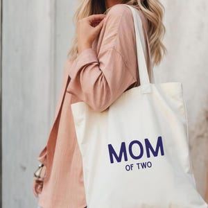 Puede incluir: Un bolso de tela color crema con el texto MOM OF TWO impreso en letras may&uacute;sculas de color azul oscuro. El bolso tiene dos correas para el hombro y una forma rectangular, confeccionado en tela tejida lisa con costuras visibles.
