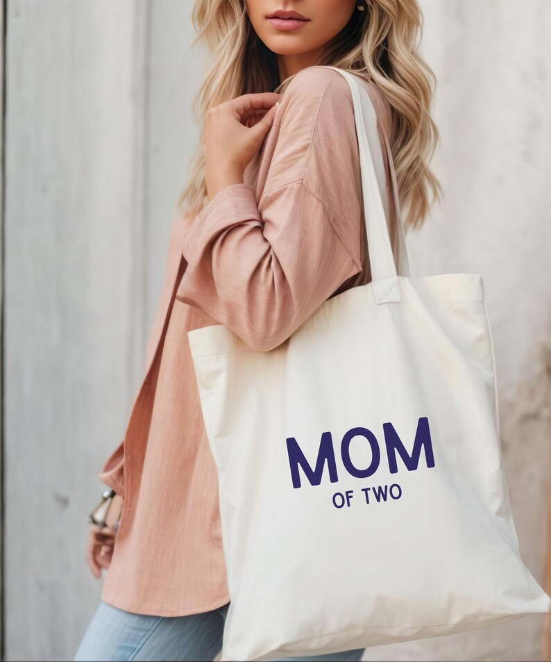 Puede incluir: Un bolso de tela color crema con el texto MOM OF TWO impreso en letras may&uacute;sculas de color azul oscuro. El bolso tiene dos correas para el hombro y una forma rectangular, confeccionado en tela tejida lisa con costuras visibles.