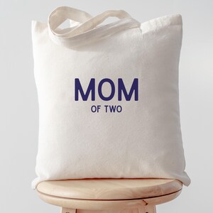 Puede incluir: Un bolso de tela color crema con dos asas superiores. La parte frontal del bolso presenta el texto "MOM OF TWO" impreso en una fuente sans-serif de color azul oscuro, con la palabra "MOM" situada sobre el texto m&aacute;s peque&ntilde;o "OF TWO".