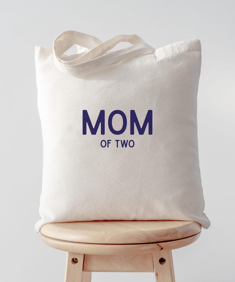 Puede incluir: Un bolso de tela color crema con dos asas superiores. La parte frontal del bolso presenta el texto "MOM OF TWO" impreso en una fuente sans-serif de color azul oscuro, con la palabra "MOM" situada sobre el texto m&aacute;s peque&ntilde;o "OF TWO".