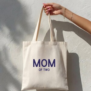 Puede incluir: Un bolso de tela de color crema con dos correas para el hombro. La parte frontal del bolso presenta el texto "MOM OF TWO" impreso en una fuente sans-serif de color azul oscuro. El bolso tiene una forma rectangular simple y bordes cosidos.