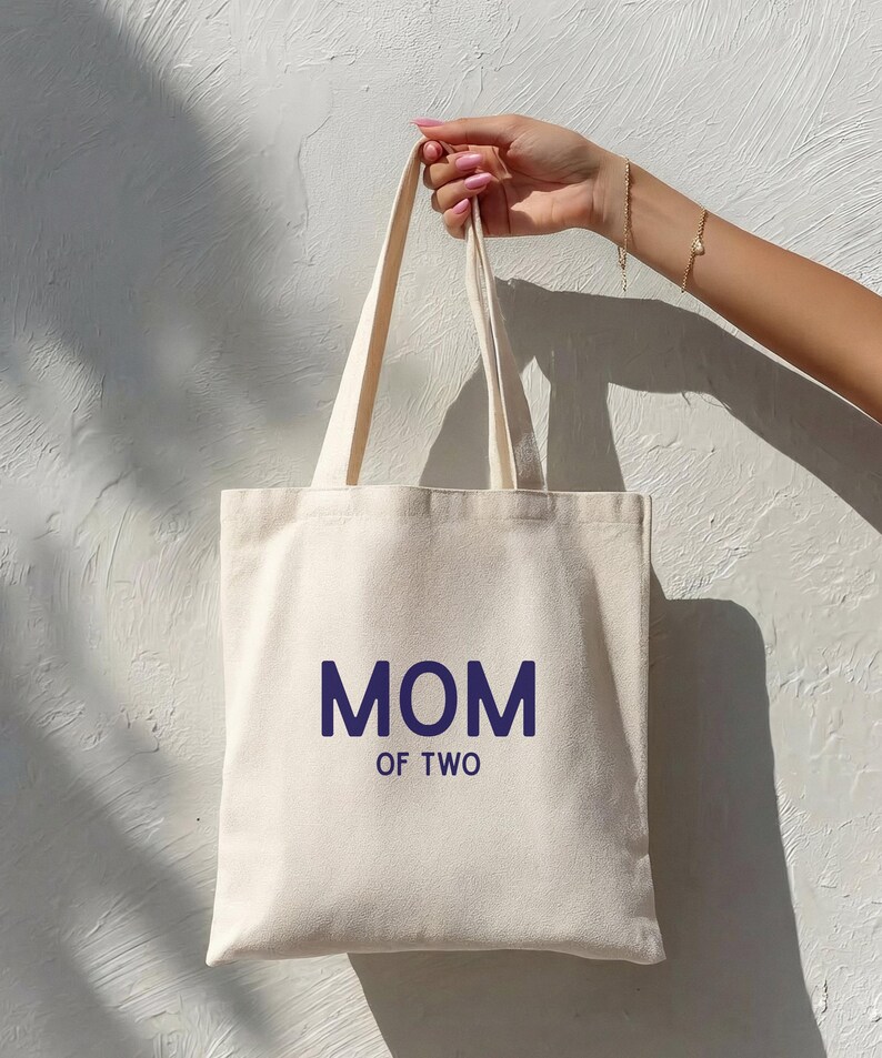 Puede incluir: Un bolso de tela de color crema con dos correas para el hombro. La parte frontal del bolso presenta el texto "MOM OF TWO" impreso en una fuente sans-serif de color azul oscuro. El bolso tiene una forma rectangular simple y bordes cosidos.
