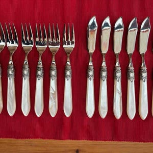 Könnte beinhalten: Ein Satz antiker Silberbestecke, darunter Gabeln und Buttermesser, auf einer roten Tischdecke angeordnet. Die Gabeln und Messer haben verzierte Silbergriffe mit Perlmutt-Finish, was dem Essgeschirr einen Hauch von Eleganz verleiht.