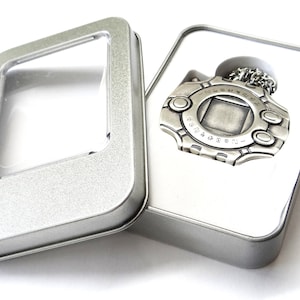 Op de afbeelding: Een zilveren Digimon-slinger met een ketting, in een zilveren blikken doosje met een transparant plastic venster.