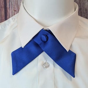 May include: A close-up of a blue cross tie, fastened with a matching button, worn over a white dress shirt. The tie's vibrant color contrasts with the shirt, creating a striking visual. The button is centered on the tie's intersection.