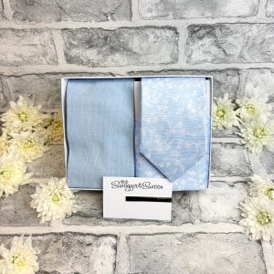 May include: A light blue tie and pocket square set with a white floral pattern, presented in a white box. The set includes a matching pocket square and tie. The box also contains a white card with the text "Swagger & Swoon".
