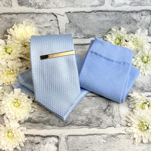 Peut inclure: Une cravate bleu clair avec un motif texturé et une pince à cravate dorée. Une pochette assortie et une paire de chaussettes bleu clair sont également présentes. Les articles sont disposés sur un fond de briques grises, avec des fleurs blanches.