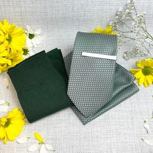 May include: A green tie, pocket square, and tie clip set with a geometric pattern. The tie clip is silver. A pair of dark green socks are also included. Yellow and white flowers are arranged around the set.