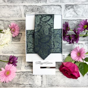 Può includere: Un set cravatta e pochette verde scuro con motivo paisley in una scatola bianca. Il set include un fermacravatta argentato e una carta con il testo "Swagger & Swoon". Lo sfondo presenta un muro di mattoni e fiori colorati.