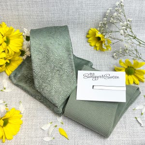 Può includere: Set cravatta e pochette verde salvia con motivo paisley e fermacravatta argentato. Il set è esposto con un biglietto bianco con la scritta "Swagger & Swoon" e fiori gialli.