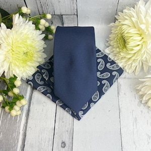 Könnte beinhalten: Eine einfarbige marineblaue Krawatte, die über einem marineblauen Einstecktuch mit weißem Paisley-Muster liegt. Das Set ist mit weißen Chrysanthemen und kleinen weißen Beeren auf einer hellgrauen Holzoberfläche arrangiert.