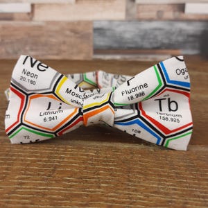 Può includere: Un papillon bianco con un design della tavola periodica, che mostra elementi come Neon, Litio e Terbio. I motivi esagonali sono delineati in colori come rosso, blu, verde e arancione. Il papillon è su una superficie di legno.