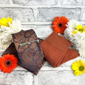 May include: A navy blue paisley tie with a silver tie clip, a matching pocket square, and a pair of rust-coloured socks. The items are arranged with orange and yellow flowers against a grey brick background.