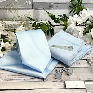Könnte beinhalten: Ein hellblaues formelles Accessoire-Set, bestehend aus Krawatte, Einstecktuch, Socken, Krawattenklammer und Manschettenknöpfen. Das Set ist auf einer Holzoberfläche mit weißen Blumen und Grünzeug im Hintergrund angeordnet. Die Manschettenknöpfe haben einen hellblauen Mittelteil.