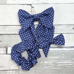 Op de afbeelding: Een verzameling marineblauwe haaraccessoires met witte stippen, waaronder twee grote strikken en een scrunchie. De strikken hebben lange uiteinden en zijn in het midden geknoopt. De scrunchie is cirkelvormig. De accessoires liggen op een lichtgrijze houten ondergrond.