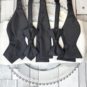 May include: Several black bow ties and neckties are arranged on a white patterned surface. The bow ties are pre-tied, and the neckties are straight. The image presents a variety of formal wear options, suitable for special occasions.