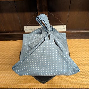 以下が含まれることがあります： 青と白のドット柄の布バッグで、上部で折りたたんで結ばれています。バッグは黒い表面に置かれ、下に黄色の織りマットがあります。上の小さな看板には「Bless this home & laughter」と書かれています。