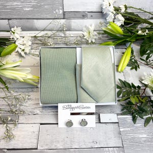 以下が含まれることがあります： 白箱に入ったセージグリーンのネクタイ、ポケットチーフ、カフスボタンのセット。セットにはネクタイ、ポケットチーフ、丸いカフスボタン2個が含まれています。「Swagger & Swoon」のブランド名がカードに印刷されています。背景には白い花があります。