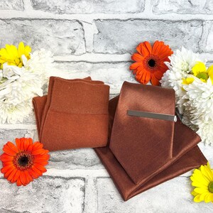 May include: A set of brown accessories including a tie with a silver clip, pocket square, and socks. The items are arranged with orange and yellow flowers against a brick wall.