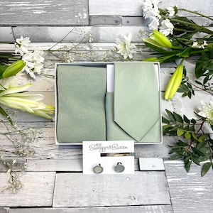 Può includere: Un set cravatta, pochette e gemelli verde salvia in una scatola bianca. Il set include un fermacravatta e due gemelli rotondi. La scatola è su una superficie di legno con fiori bianchi e verde.
