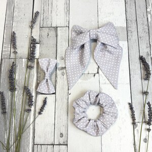 Pode incluir: Um conjunto de três acessórios de cabelo: um laço grande, um laço pequeno e uma scrunchie. Todos são cinza claro com bolinhas brancas. Os itens estão dispostos sobre uma superfície de madeira cinza clara desgastada, com raminhos de lavanda. A scrunchie tem cerca de 10 cm de diâmetro.