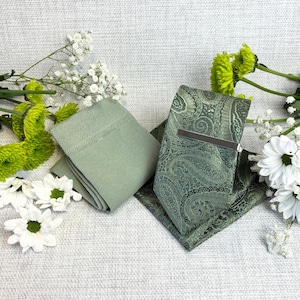 May include: A sage green tie, pocket square, and socks set with a paisley pattern. The tie has a silver tie clip. The set is displayed with white and green flowers on a textured, light grey surface.