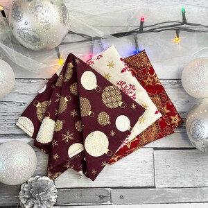 Op de afbeelding: Vier kerstservetten in bordeauxrood, crème en rood, met gouden accenten. De servetten tonen feestelijke patronen van ornamenten, sneeuwvlokken en sterren. Ze zijn gerangschikt op een licht houten oppervlak.