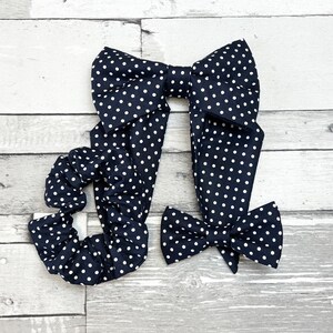 Puede incluir: Un conjunto de accesorios para el cabello con lunares azul marino, que incluye un lazo grande, un lazo más pequeño y una goma para el pelo. Los lunares blancos están espaciados uniformemente sobre la tela. Los artículos están colocados sobre una superficie de madera clara.