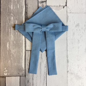 May include: A dusty blue bow tie with two long, rectangular fabric tails hanging downwards, positioned over a matching square pocket square. The set features a smooth, solid-coloured fabric with a visible weave and a small metal hook fastener.