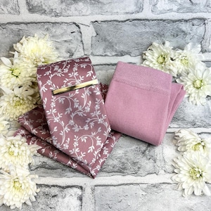 May include: A mauve tie and pocket square set with a white floral design and a gold tie clip. Beside the set are mauve socks. The items are arranged on a grey brick background with white flowers.