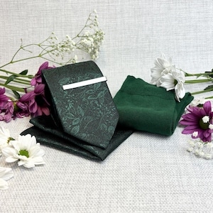 Pode incluir: Uma gravata verde escuro com um padrão de folhas e um prendedor de gravata prateado, um lenço de bolso a condizer e um par de meias verdes. Os artigos estão dispostos com flores brancas e roxas numa superfície texturizada.