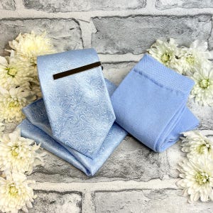 May include: A light blue paisley tie, pocket square, and socks set. The tie features a silver tie clip. The items are displayed on a grey brick background, decorated with white flowers.