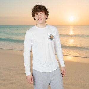 May include: White long-sleeve shirt with a crew neck and a small circular graphic on the left chest. The graphic features a beach scene with the words "Hooked Daily" in a circular design. The shirt is paired with gray sweatpants.
