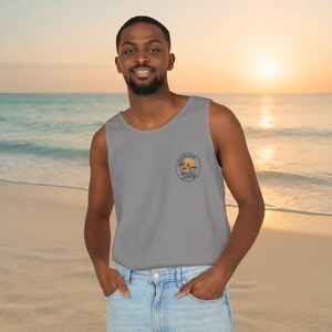 May include: A gray tank top with a round embroidered patch featuring a palm tree, sun, and the text "Hooked & Salty". The tank top is worn by a person on a beach with the ocean and sunset in the background.