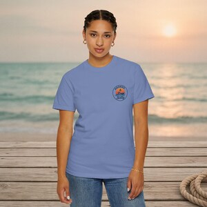 May include: A periwinkle blue short-sleeve t-shirt featuring a circular graphic on the left chest. The graphic depicts a sunset over the ocean with a boat silhouette and the text Hooked & Salty written in a dark blue circular border.