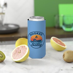 May include: A light blue can cooler with the words "Hooked & Salty" in a circular design featuring a boat on the water at sunset. The cooler is holding a silver can. Grapefruit and kiwi are in the foreground.